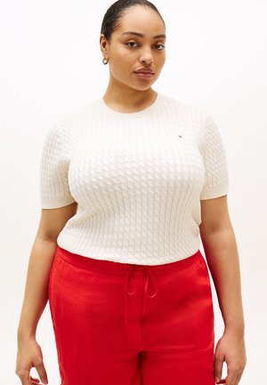 Žena koja nosi bijeli teksturirani džemper kratkih rukava i crvene hlače s vezicom, stoji ispred jednobojne pozadine.