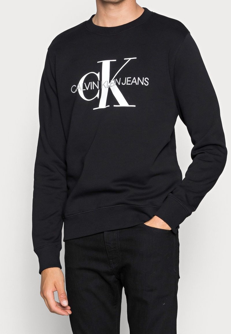 Homme portant un sweat Calvin Klein Jeans noir avec un grand logo blanc "CK" et texte, pantalon noir, une main dans la poche.