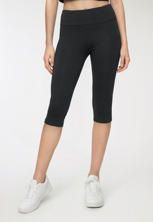 Donna con leggings neri corti e sneakers bianche, in piedi contro uno sfondo chiaro e uniforme.