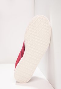 Röd mockaskor med en texturerad vit gummisula. Synlig sömnad längs sidan och en rundad, låg design.