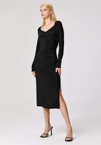 Femme aux longs cheveux blonds portant une robe midi noire à manches longues avec une fente sur le côté et des sandales à talons noirs avec des brides en strass.