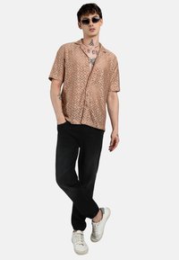Camicia a maniche corte beige con motivo a diamante, abbinata a pantaloni neri e sneakers bianche. Gli accessori includono occhiali da sole.