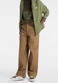 Khaki wijde broek met een sluiting aan de voorkant, gecombineerd met een groene hoodie met rits en een gestreept groen sweater. Bruine en witte sneakers.