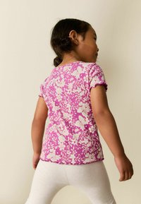 Lila virágos, rövid ujjú póló texturált mintával és fodros szegéllyel, fehér legginggel párosítva. A póló hátulnézete látható.