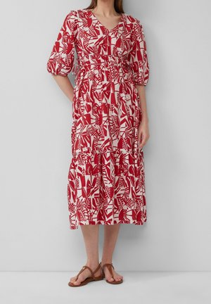 Femme portant une robe midi à motifs rouges et blancs avec des manches bouffantes et des sandales plates marron, debout devant un fond clair uni.