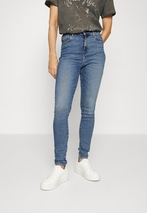 Personne portant un jean skinny bleu délavé, des baskets blanches compensées et un t-shirt graphique foncé avec un motif tigre.