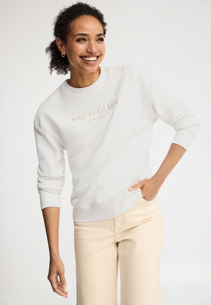 Donna sorridente che indossa una felpa bianca con la scritta "Polo Club" e pantaloni beige chiaro con una mano in tasca, su sfondo semplice.