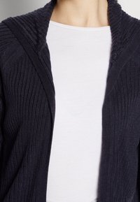 Námořnický cardigan s žebrovanou strukturou a šálovým límcem, nošený přes jednobarevné bílé tričko. Má těsný střih a měkkou látku.