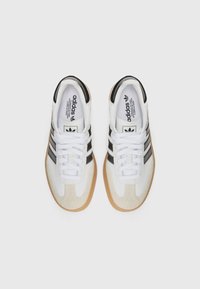 Zapatillas deportivas blancas con parte superior de ante y sintético, diseño de tres rayas negras y suela de goma beige. Los cordones son blancos.