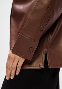 Casaco de couro castanho com um punho largo com fecho de botão de pressão e uma fenda lateral na bainha, apresentando uma textura suave.