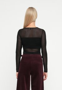 Donna con capelli biondi lisci che indossa un top trasparente nero a maniche lunghe e pantaloni di velluto a coste viola scuro a vita alta, voltata di spalle rispetto alla fotocamera.