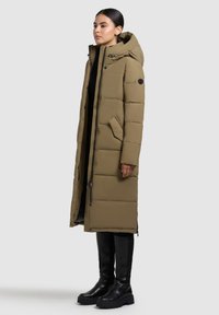 Parka lunga color verde oliva con cappuccio, chiusura a zip frontale e tasche laterali. Presenta una texture trapuntata e una silhouette aderente, abbinata a stivali neri al ginocchio.
