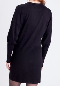 Abito nero lavorato a maglia con maniche a sbuffo e gonna aderente a costine, texture liscia e scollatura a girocollo. Il modello è mostrato di spalle.