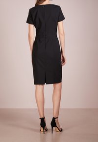 Robe noire cintrée avec manches courtes et ourlet à hauteur des genoux. Comprend une fente au dos et une fermeture ronde au niveau du cou. Texture de tissu lisse.