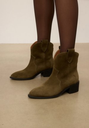 Bottines en daim marron à petits talons carrés portées avec des collants noirs transparents sur un sol beige.