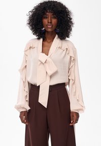 Beżowa bluzka z falbanastymi rękawami i kokardą na szyi, zestawiona z brązowymi spodniami o wysokim stanie. Miękka, gładka tekstura tkaniny.