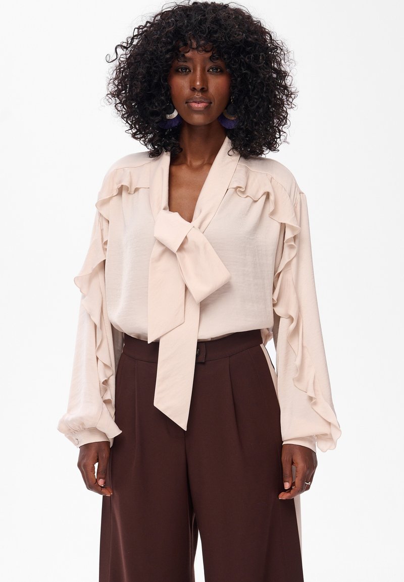 Beżowa bluzka z falbanastymi rękawami i kokardą na szyi, zestawiona z brązowymi spodniami o wysokim stanie. Miękka, gładka tekstura tkaniny.
