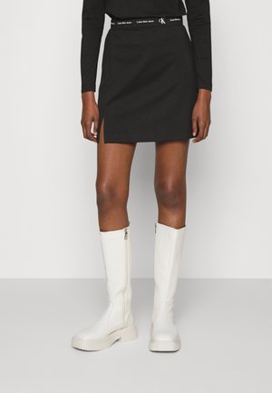 Minifalda negra con abertura lateral y cintura con logotipo, combinada con botas blancas hasta la rodilla con suela texturizada y cierre de cremallera.