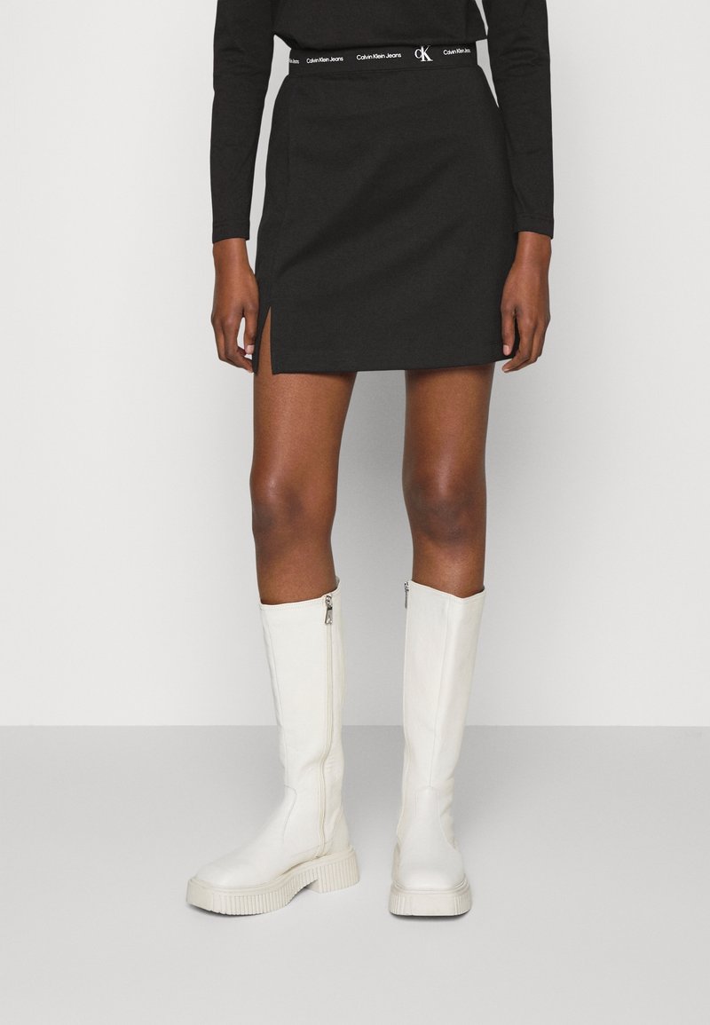 Black mini skirt with side slit and logo waistband, paired with knee-high white boots featuring a textured sole and zip closure.