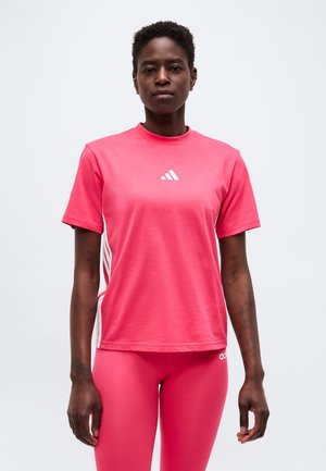 Kvinde iført pink Adidas t-shirt og leggings, står mod en ensfarvet lys baggrund, vendt fremad med afslappede arme.