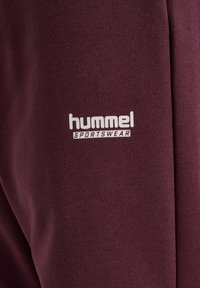 Close-up van bordeauxrode stof met wit "hummel SPORTSWEAR" logo erop gedrukt.