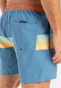 Bañador azul con una cintura elástica marrón, que presenta rayas verticales multicolores (beige, azul claro, amarillo) y un bolsillo trasero con un botón a presión.