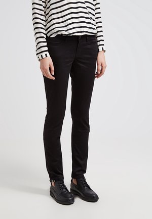 Person iført sorte smalle bukser, en langærmet hvid og sortstribet skjorte og sorte snørestøvler på en hvid baggrund.