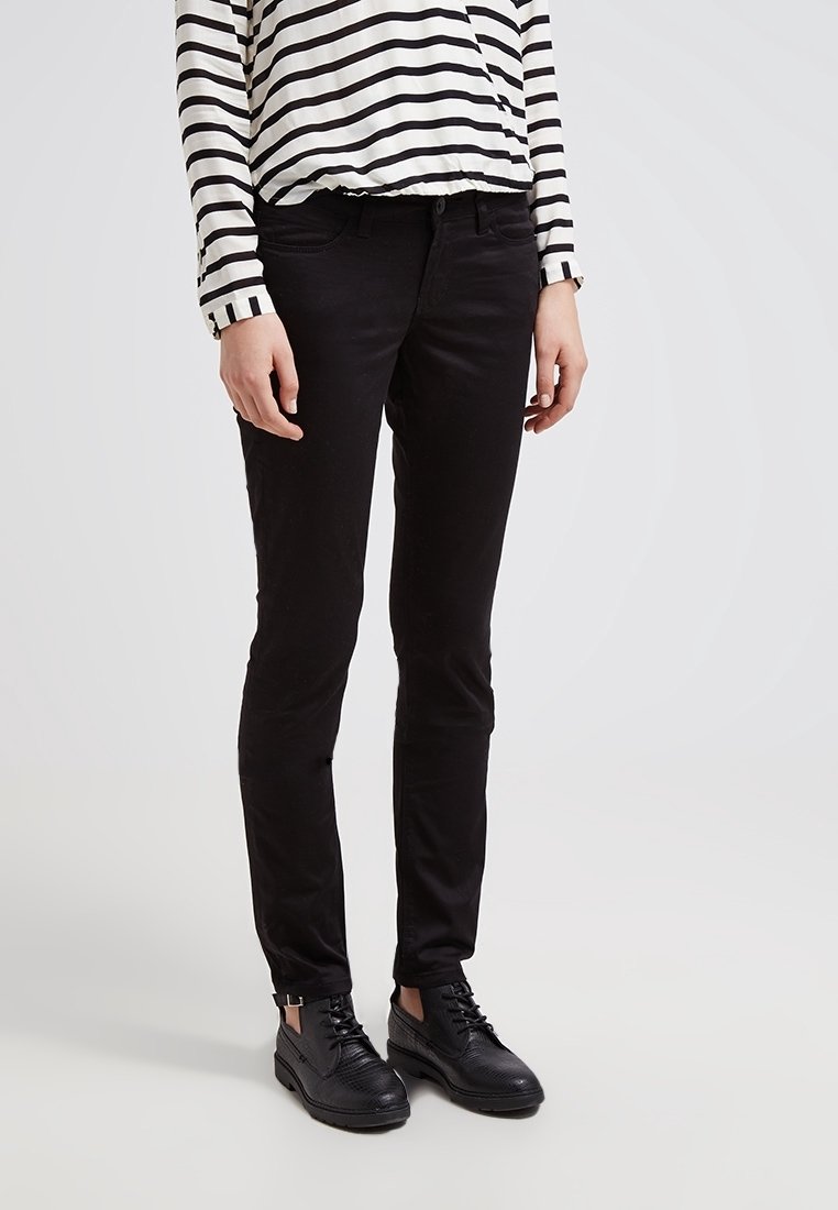 Personne portant un pantalon noir ajusté, une chemise à manches longues rayée blanche et noire, et des chaussures noires à lacets sur un fond blanc.