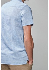 Chemise bleu clair à manches courtes avec un tissu texturé, dotée d'un col pointu et d'une coupe décontractée, rentrée dans un pantalon kaki.