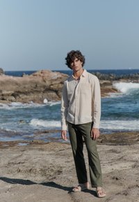 Chemise à manches longues beige clair, pantalon vert olive et sandales légères. Arrière-plan naturel avec des rochers et l'océan, ciel bleu clair.