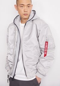 Veste bomber argentée avec des poignets et un ourlet côtelés, dotée d'une capuche grise, d'une fermeture éclair à l'avant et d'une étiquette rouge sur la manche. Texture lisse.