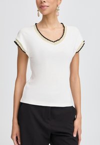 Top bianco a maniche corte con scollatura a V, caratterizzato da un bordo nero a scallop e un accento dorato lungo il collo e le maniche. Tessuto a maglia liscia.