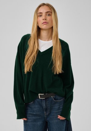 Ung kvinde med langt blondt hår iført en mørkegrøn V-hals sweater, hvid top under og blå jeans med sort bælte.
