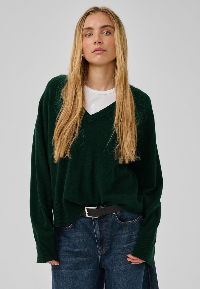 Jeune femme aux longs cheveux blonds portant un pull vert foncé à col en V, un top blanc en dessous, et un jean bleu avec une ceinture noire.