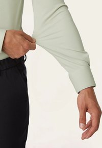 Lichtgroene langekoortriem met een fitted manchette, die bij de pols wordt uitgerekt. De stof lijkt glad en iets rekbaar.