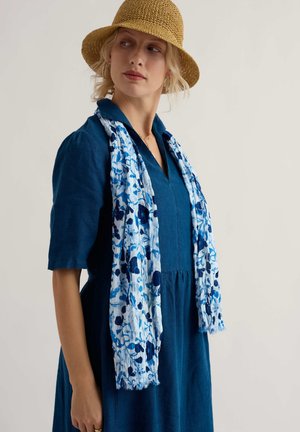 Žena v modrých šatách, svetlomodrom kvetovanom šále a béžovom pletenom klobúku, pozerajúca sa bokom na jednoliatom pozadí.