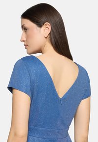 Femme aux longs cheveux bruns raides portant une robe bleue scintillante avec un dos en V profond et des manches courtes, tournée vers la gauche.