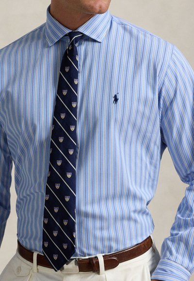 Mann trägt hellblau gestreiftes Hemd mit marineblauer gemusterter Krawatte und braunem Ledergürtel, in weiße Hose gesteckt.