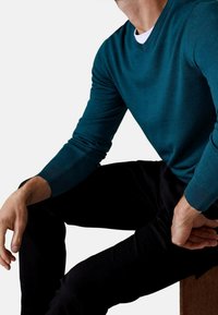 Maglione lavorato a maglia color teal con scollo a V, maniche lunghe, polsini a costine e una texture liscia, abbinato a pantaloni neri.