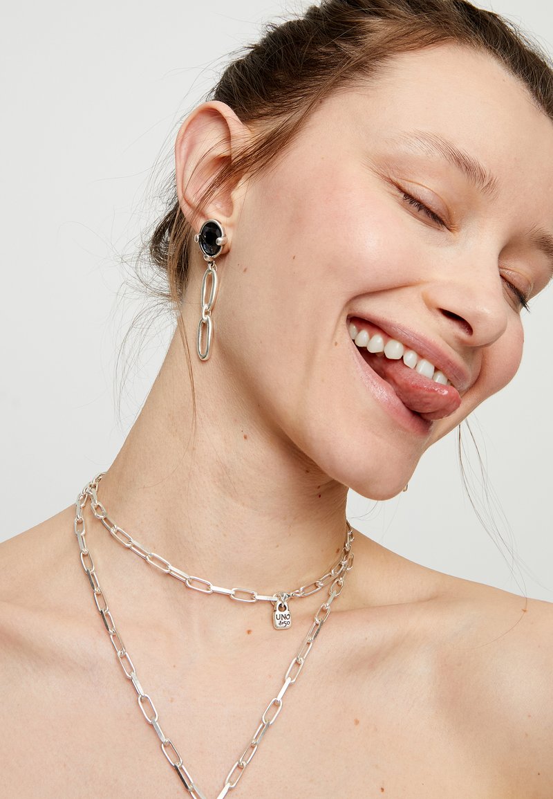 Collar de cadena de plata con eslabones; diseño en capas. Pendientes colgantes con una piedra negra y detalles ovalados de plata. Tono de piel claro.