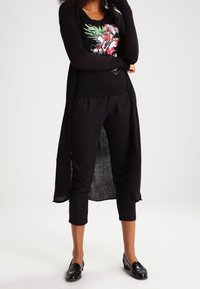 Černý dlouhý cardigan přes černé grafické tričko s květinovým designem. Černé cropped kalhoty a lesklé černé mokasíny dotvářejí celkový vzhled.