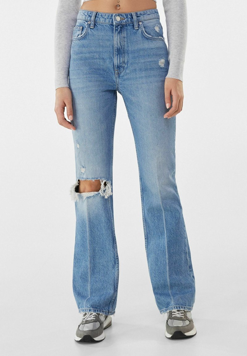 Högmidjade, ljusblå slitna jeans med fransade knäöppningar och en lätt utvidgning, med en traditionell femfickorsdesign.