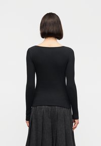 Personne aux cheveux courts et foncés, portant un haut noir côtelé à manches longues ajusté et une jupe plissée gris foncé, vue de dos sur un fond uni.