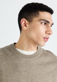 Pull en maille marron clair avec un col rond, porté sur une chemise crème. Le mannequin a les cheveux courts et texturés et porte une petite boucle d'oreille en or.