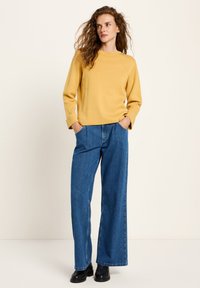 Frau mit lockigem Haar, die einen senfgelben Pullover, weite blaue Jeans und schwarze Schuhe trägt und mit den Händen in den Taschen steht.