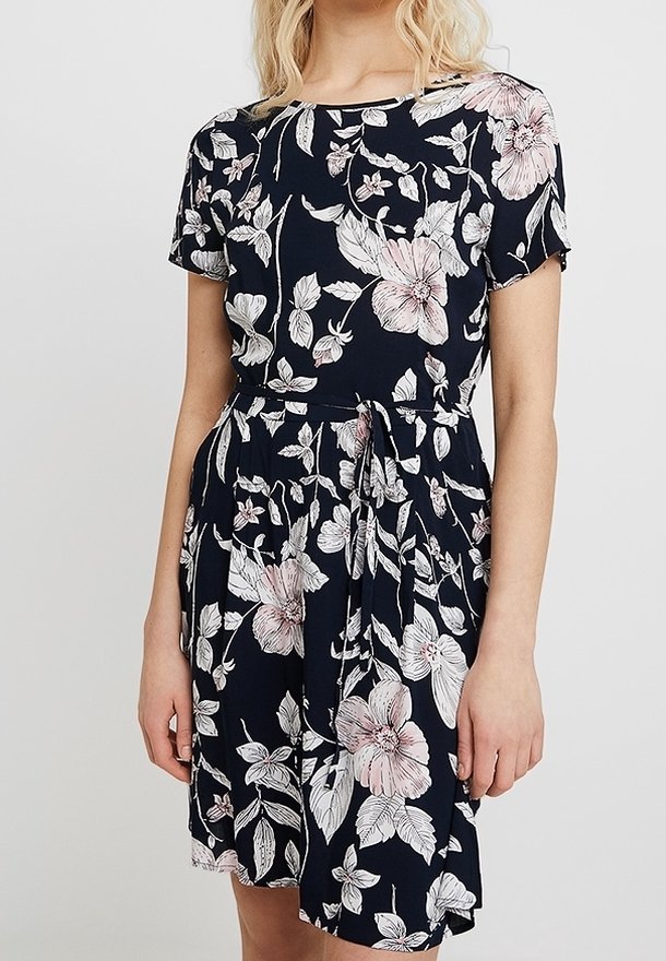 Femme portant une robe noire avec un imprimé floral rose clair et blanc, manches courtes et ceinture nouée à la taille.