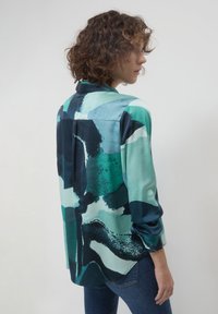 Frau mit lockigem Haar, die eine blau-grün gemusterte Bluse und blaue Jeans trägt, seitlich vor einem schlichten weißen Hintergrund stehend.
