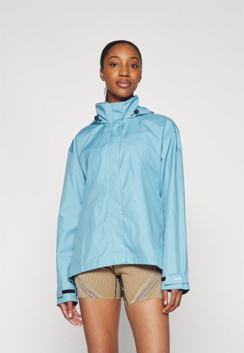 Giacca impermeabile azzurra chiara con colletto alto e cappuccio regolabile. Presenta chiusura con zip e polsini elasticizzati. Abbinata a pantaloni corti a righe.
