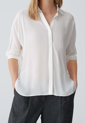 Femme portant un chemisier blanc ample à boutons avec manches retroussées et pantalon gris foncé texturé, mains dans les poches, sur un fond uni.