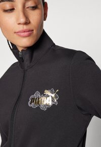 Giacca nera con zip caratterizzata da un design floreale e logo metallico dorato. Texture morbida con colletto alto e forma aderente.